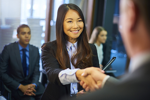 young woman steps forward as she is called in for her interview and shakes hands with her possible new employer. Behind her other candidates wait with their resumes.